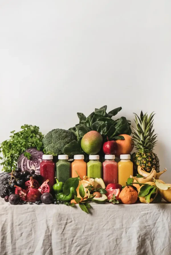 Sechs bunte Flaschen mit Saft sind in einer Reihe zwischen einer bunten Auswahl an frischem Obst und Gemüse wie Spinat, Brokkoli, Ananas, Mango und Trauben auf einer hellen Tischdecke vor einem weißen Hintergrund ausgestellt