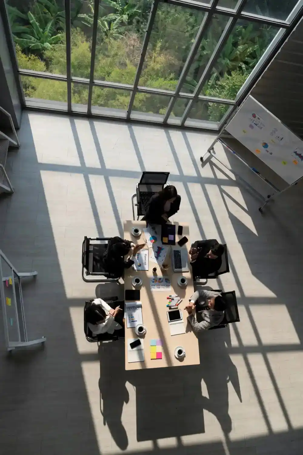 Draufsicht auf die Fachkräfte der Gesundheits Zone, die in einem sonnendurchfluteten Büroraum mit großer Glasdecke und Blick ins Grüne an einem hellen Holzkonferenztisch zusammenarbeiten.